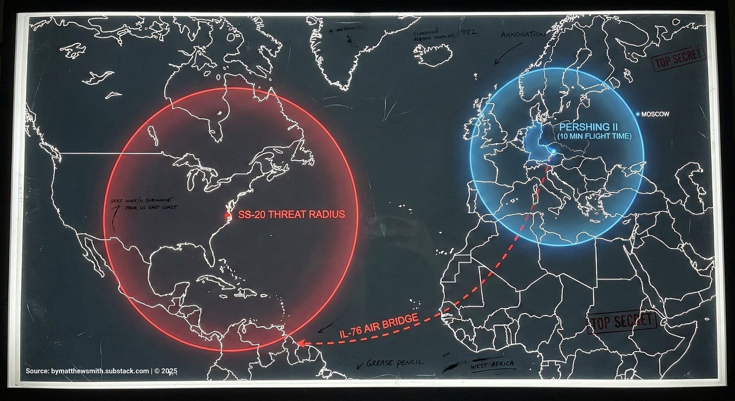 1982 Cold War tactical map visualization on a lightbox. A blue radius circle shows US Pershing II missile range extending from West Germany to Moscow. A red radius circle shows Soviet SS-20 missile range extending from Suriname to cover the entire US East Coast and Midwest. A dashed line shows an IL-76 cargo flight path from West Africa to Suriname.