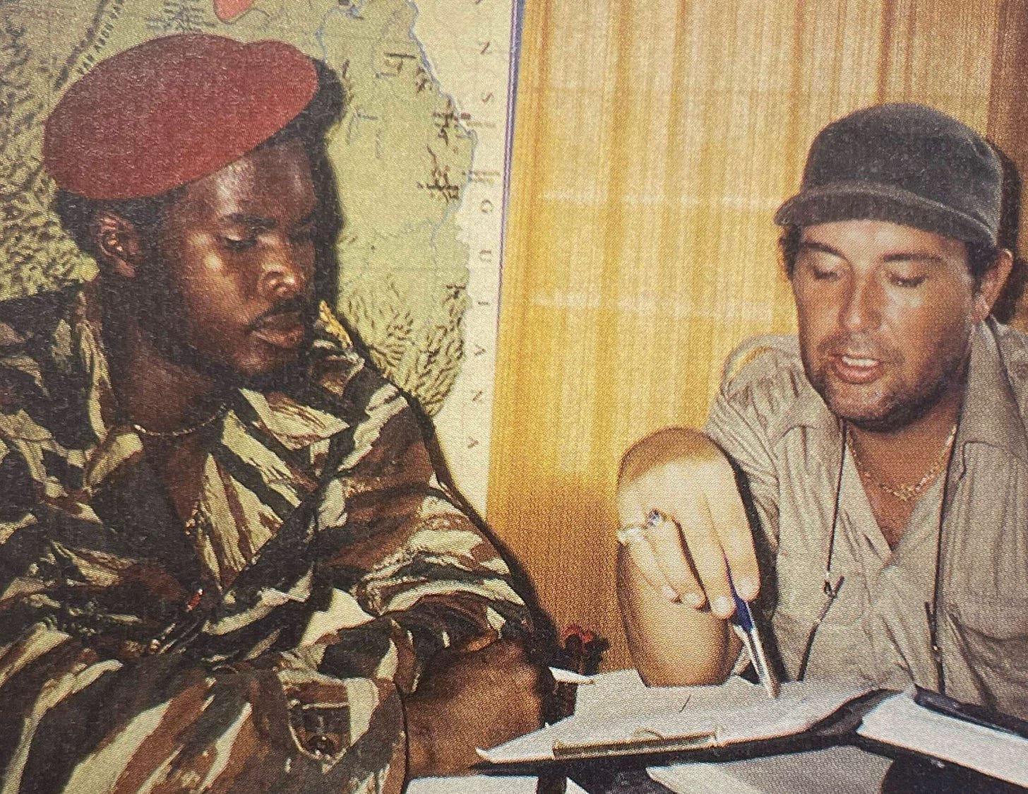 A historical photograph of Ronnie Brunswijk and Frits Hirschland sitting at a table together. Brunswijk, on the left, wears a red beret and tiger-stripe camouflage. Hirschland, on the right, wears a grey cap and a tan shirt, pointing at a document with a pen. A map of Suriname and the Guianas is visible in the background. A historical photograph of Ronnie Brunswijk and Frits Hirschland sitting at a table together. Brunswijk, on the left, wears a red beret and tiger-stripe camouflage. Hirschland, on the right, wears a grey cap and a tan shirt, pointing at a document with a pen. A map of Suriname and the Guianas is visible in the background.